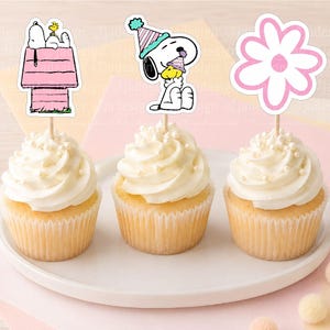 Pode incluir: Três cupcakes cobertos com glacê branco e topos de papel decorativos. Os topos apresentam o Snoopy numa casota cor-de-rosa, o Snoopy com um chapéu de festa a segurar o Woodstock e uma flor cor-de-rosa e branca em palitos de madeira.