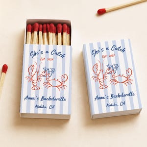 May include: Two rectangular matchboxes with light blue and white vertical stripes. The design features a red lobster and crab illustration, blue text reading She's a Catch, Est. 2027, Anna's Bachelorette, and Malibu, CA. Several matches with red tips are visible.
