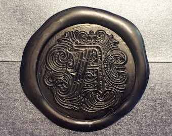 Op de afbeelding: Een zwarte waszegel met een decoratief ontwerp met een letter "A" in het midden.