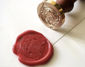 Op de afbeelding: Een rode waszegel met een bloemmotief, gedrukt met een goudkleurige stempel. De stempel heeft een gedetailleerd bloemmotief.