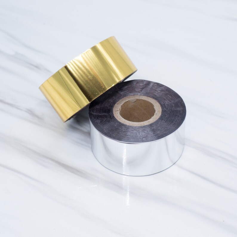 Op de afbeelding: Twee rollen folie, een gouden en een zilveren, liggen op elkaar. De rollen hebben beide een diameter van ongeveer 2,5 cm.