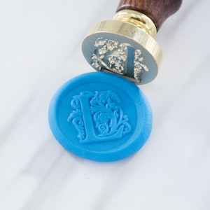 Peut inclure: Un sceau de cire bleu avec un motif décoratif en forme de lettre L. Le sceau est réalisé à l'aide d'un tampon à cire et sert à sceller des enveloppes ou des documents.