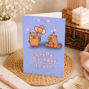 May include: A light purple birthday greeting card featuring two brown capybaras, one holding a bunch of balloons and the other wearing a party hat with a cake and gifts, with the text Cappy Birthday to you printed in pink bubble letters.