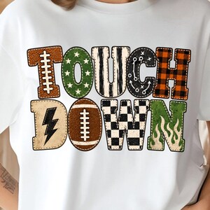 May include: A white short-sleeve t-shirt featuring the text TOUCH DOWN in large, multi-patterned letters. The design includes football textures, green stars, black and white stripes, play diagrams, orange plaid, a lightning bolt, and flames.