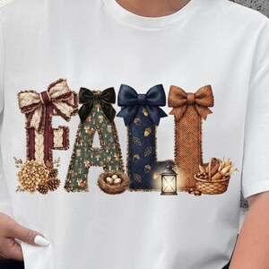 Op de afbeelding: Een wit t-shirt met korte mouwen en het woord FALL in grote letters. Elke letter is versierd met een strik en seizoensgebonden afbeeldingen, waaronder een vogelnest, een lantaarn, een mand met maïs en patronen van vossen, paddenstoelen en eikels.