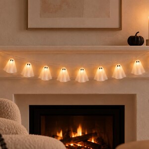 May include: A string of eleven white ghost-shaped LED lights, each featuring two small black oval eyes. The translucent ghost figures are illuminated from within, emitting a warm glow, and are connected by a thin, flexible wire cord.