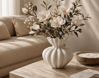 May include: A white ceramic vase with a scalloped, ribbed design holds a bouquet of white magnolia flowers and small green leafy branches. The vase has a wide, rounded base that tapers into a narrow neck before flaring out at the top rim.