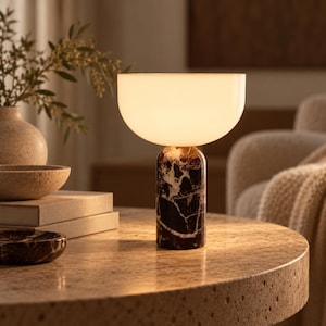 Puede incluir: Una lámpara de mesa con una base cilíndrica de mármol veteado en marrón oscuro y blanco, rematada con una gran pantalla hemisférica de vidrio blanco esmerilado que emite una luz cálida y suave al encenderse.