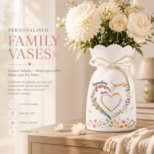 Könnte beinhalten: Eine weiße Keramikvase mit gewelltem Rand und einem cremefarbenen Band um den Hals. Die Vase zeigt ein buntes, herzförmiges Blumendesign mit dem Text "Grandma" und "Mom" sowie verschiedenen Namen und einer floralen Bordüre am unteren Rand.