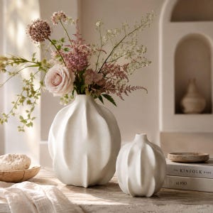 Op de afbeelding: Twee witte keramische vazen met een geribbelde, organische, pompoenachtige vorm. De grotere vaas bevat een boeket van roze rozen, paarse alliums en groen blad. De kleinere vaas staat ernaast op een oppervlak met een boek genaamd Kinfolk Home.