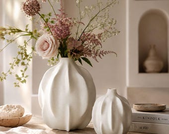 May include: Two white ceramic vases with a ribbed, organic, pumpkin-like shape. The larger vase holds a bouquet of pink roses, purple alliums, and green foliage. The smaller vase sits next to it on a surface with a book titled The Kinfolk Home.