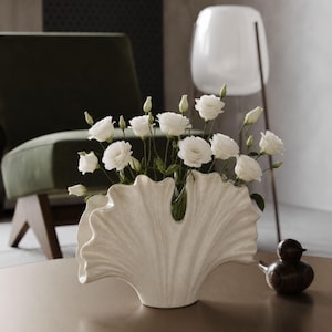 Op de afbeelding: Een crèmekleurige keramische vaas met een brede, waaiervormige structuur en een gespikkelde afwerking. De vaas bevat een boeket witte eustoma-bloemen met meerdere open bloemen en gesloten knoppen op slanke groene stelen in een waaier.
