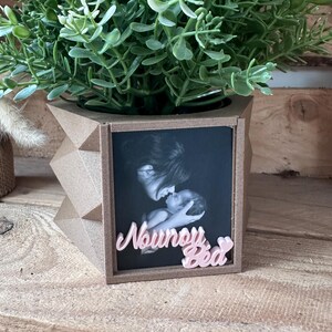 Peut inclure: Un pot de fleurs géométrique de couleur beige contenant une plante artificielle verte. L'avant du pot présente une photographie en noir et blanc d'un adulte tenant un bébé, avec le texte rose en 3D "Nounou Béa" fixé au bas du cadre.