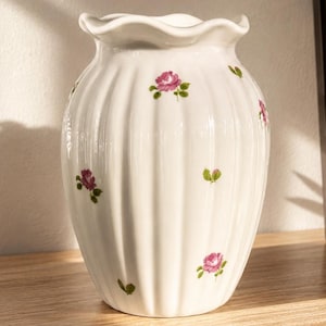 Könnte beinhalten: Weiße Keramikvase mit gerüschtem Rand und vertikalem Rippendesign. Die Vase ist mit kleinen rosa Rosen und grünen Blättern verziert. Die Vase steht auf einer Holzoberfläche.