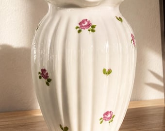 May include: White ceramic vase with a ruffled rim and vertical ribbed design. The vase is adorned with small pink rose and green leaf motifs. The vase is sitting on a wooden surface.