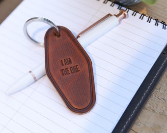 May include: A brown leather keychain with the text "I AM THE ONE" engraved on it. The keychain is in the shape of a hotel key and is attached to a silver metal ring. A white pen with rose gold accents rests on a spiral notebook.