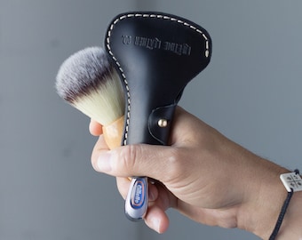May include: A black leather razor case with white stitching and the text "LIFETIME LEATHER CO." holds a shaving brush and a Gillette razor. The brush has a wooden handle and white and brown bristles.