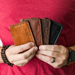 Puede incluir: Una persona sosteniendo cuatro porta tarjetas de cuero en diferentes colores. Los porta tarjetas est&aacute;n hechos por "Genuine Leather Co."