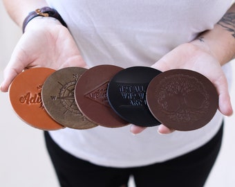 May include: A set of five circular leather coasters in varying shades of brown and black. Each coaster features a unique embossed design, including a compass, a tree, and text. The coasters are arranged in a fan shape, displaying different textures.
