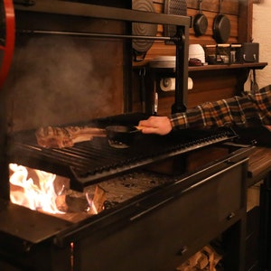 May include: A black metal grill with a large piece of meat cooking over an open fire. The grill has a raised grate and a lower section for the fire. A person is seen tending to the grill.