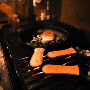 May include: Three cast iron skillets with leather handles on a grill. The skillets are on a black grill grate with glowing embers underneath. One skillet has a cooked steak and vegetables inside. The other two skillets have garlic cloves inside.
