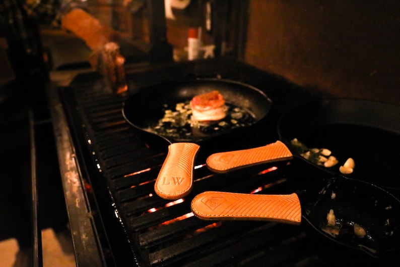 May include: Three cast iron skillets with leather handles on a grill. The skillets are on a black grill grate with glowing embers underneath. One skillet has a cooked steak and vegetables inside. The other two skillets have garlic cloves inside.