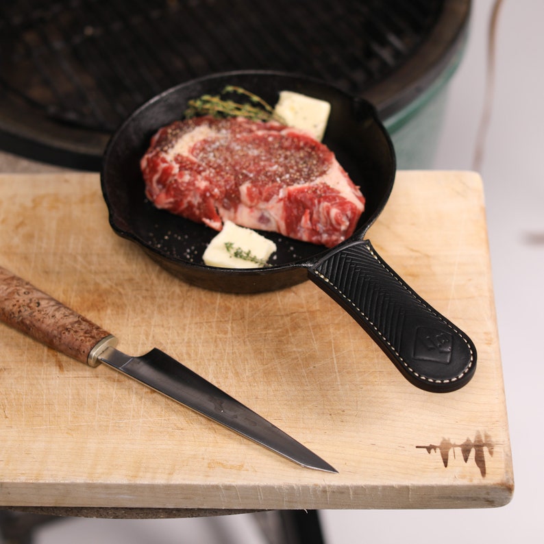 May include: A black cast iron skillet with a leather handle, containing a raw steak with butter and herbs. A wooden cutting board with a knife is in the foreground.