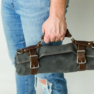 May include: A gray canvas roll bag with brown leather straps and silver buckles. The bag has a handle and is held by a person wearing blue jeans. The bag is designed to hold tools or other items.
