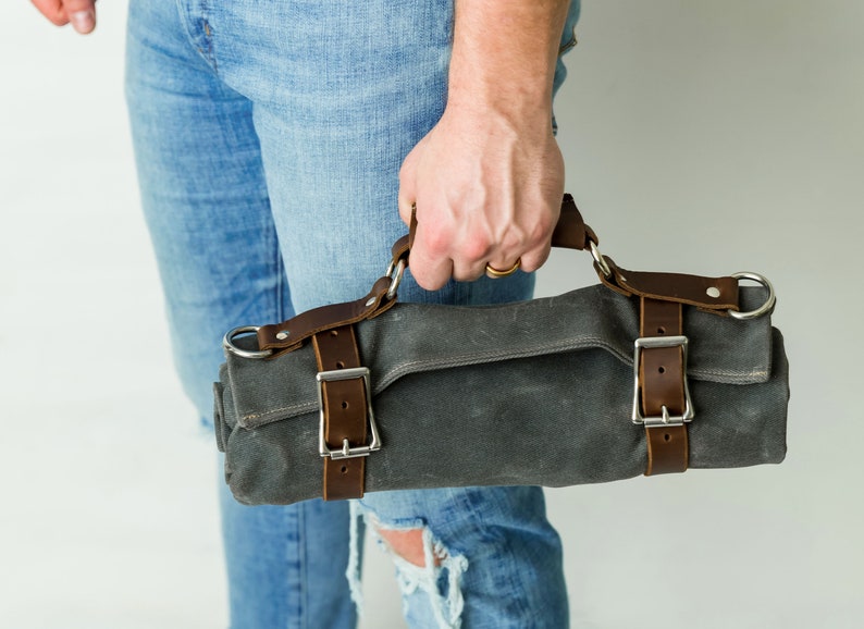 May include: A gray canvas roll bag with brown leather straps and silver buckles. The bag has a handle and is held by a person wearing blue jeans. The bag is designed to hold tools or other items.
