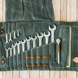 May include: A gray canvas tool roll with a collection of wrenches and a ratchet. The roll is open, displaying the tools in individual pockets. Brown leather straps and accents secure the roll. The background is a light wood surface.