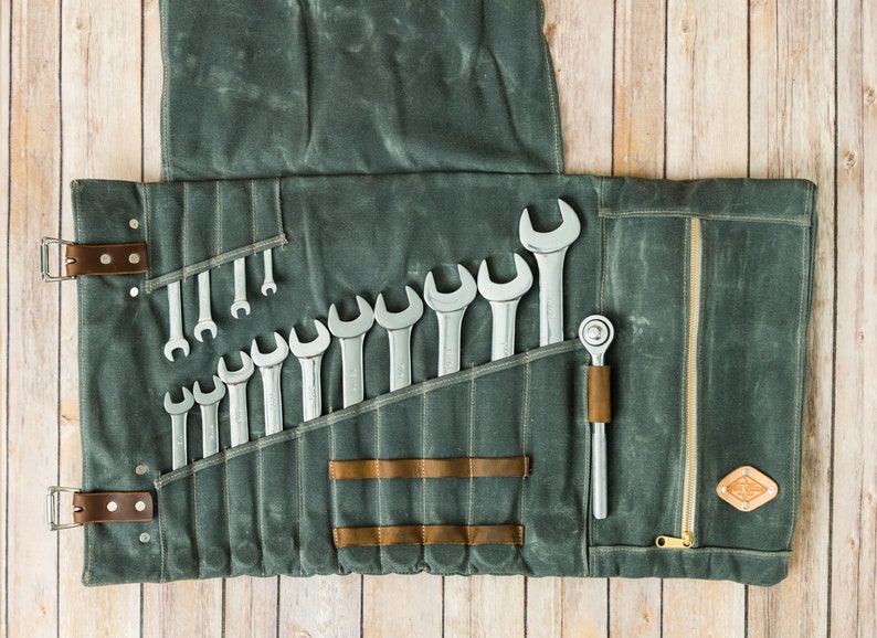 May include: A gray canvas tool roll with a collection of wrenches and a ratchet. The roll is open, displaying the tools in individual pockets. Brown leather straps and accents secure the roll. The background is a light wood surface.