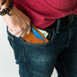 Puede incluir: Una persona con jeans azules y una camiseta roja sostiene una cartera de cuero marr&oacute;n con una tarjeta de cr&eacute;dito azul y dinero en efectivo en su interior. La cartera tiene el texto "Craft Leather" grabado en relieve.