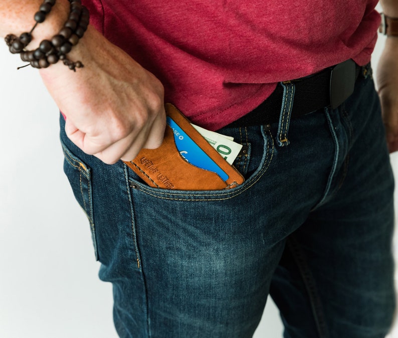 Puede incluir: Una persona con jeans azules y una camiseta roja sostiene una cartera de cuero marr&oacute;n con una tarjeta de cr&eacute;dito azul y dinero en efectivo en su interior. La cartera tiene el texto "Craft Leather" grabado en relieve.