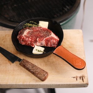 May include: A seasoned ribeye steak in a cast iron skillet with a leather handle. The skillet is on a wooden cutting board with a small knife next to it.