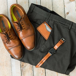 Puede incluir: Un par de zapatos marrones de cuero con cordones, una cartera de cuero marr&oacute;n y una correa de reloj de cuero marr&oacute;n en un pantal&oacute;n de vestir gris.