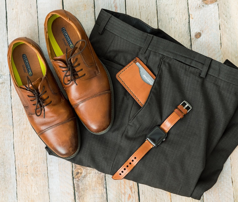 Puede incluir: Un par de zapatos marrones de cuero con cordones, una cartera de cuero marr&oacute;n y una correa de reloj de cuero marr&oacute;n en un pantal&oacute;n de vestir gris.