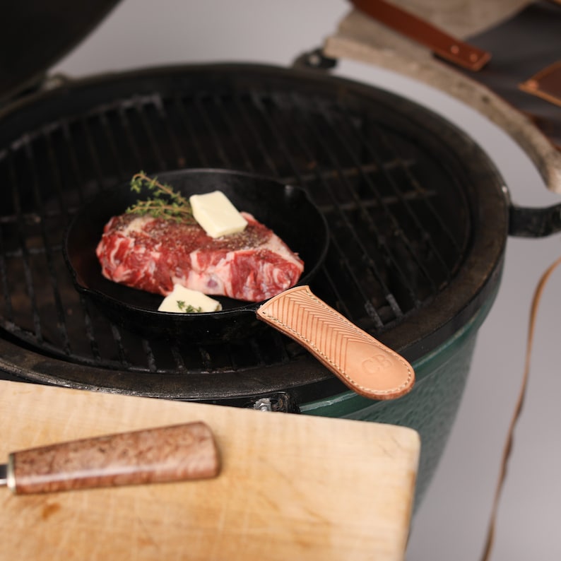 May include: A cast iron skillet with a leather handle sits on a green grill. Inside the skillet is a raw steak with butter and thyme.
