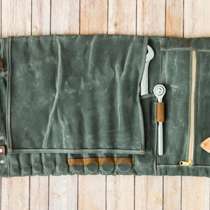 May include: A gray canvas tool roll with leather accents, open and laid flat on a wooden surface. The roll contains a wrench and a ratchet. The roll has a zipper closure and leather straps with buckles.