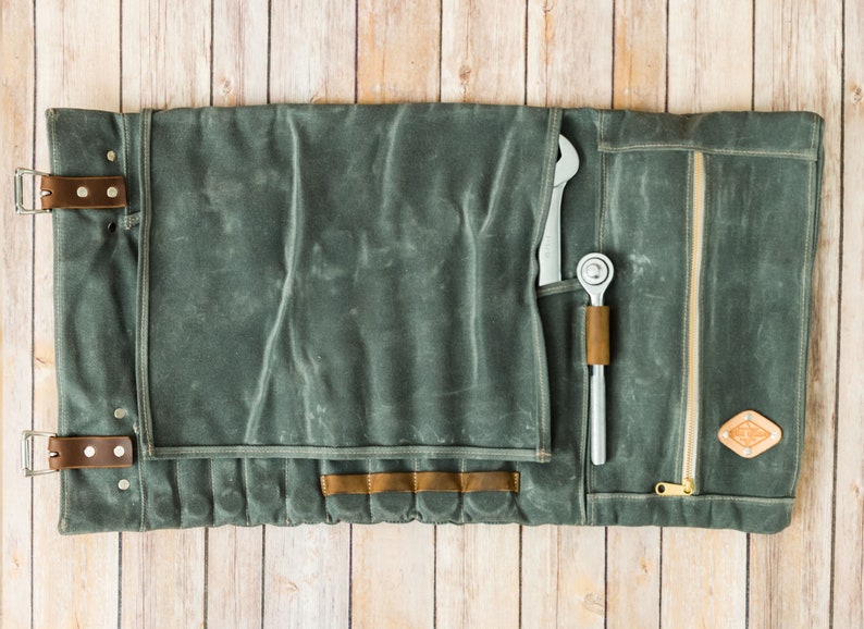 May include: A gray canvas tool roll with leather accents, open and laid flat on a wooden surface. The roll contains a wrench and a ratchet. The roll has a zipper closure and leather straps with buckles.