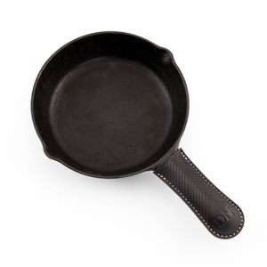 May include: A black cast iron skillet with a leather handle. The handle has a chevron pattern and the initials "DN" are embossed on it.