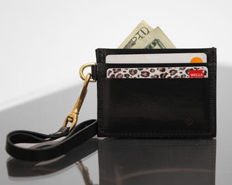 May include: A black leather card holder with multiple slots containing a twenty-dollar bill and two credit cards, one featuring a leopard print design. A gold-toned metal lobster clasp attaches a black leather wrist strap to the side of the holder.