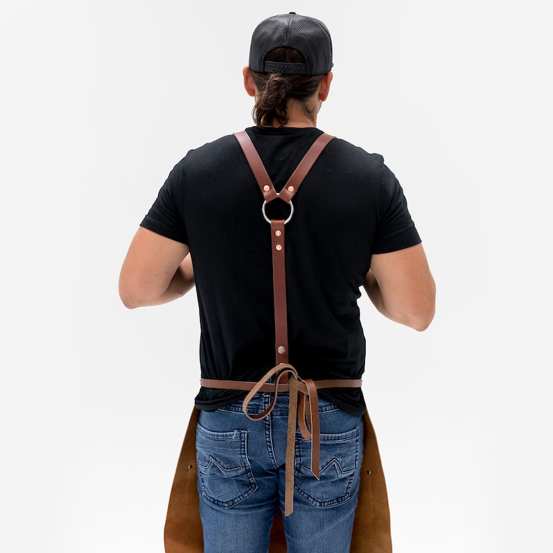 May include: Brown leather apron with suspenders, featuring a large metal ring at the back and a tie at the waist. The apron is worn over a black shirt and blue jeans. The leather has a natural, textured appearance.