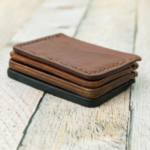 Puede incluir: Tres carteras de cuero apiladas una encima de la otra. La cartera superior es de cuero marr&oacute;n con un borde cosido. La cartera del medio es de cuero marr&oacute;n y la cartera inferior es de cuero negro.