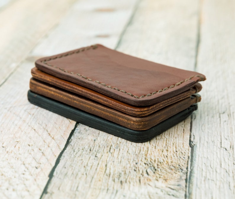 Puede incluir: Tres carteras de cuero apiladas una encima de la otra. La cartera superior es de cuero marr&oacute;n con un borde cosido. La cartera del medio es de cuero marr&oacute;n y la cartera inferior es de cuero negro.