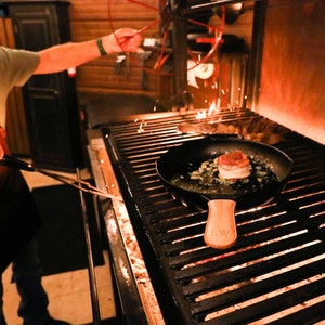 May include: A close-up of a cast iron skillet with a wooden handle, sitting on a black grill. The skillet contains a cooked steak with a bacon wrap and green onions. The grill is lit and has flames visible.