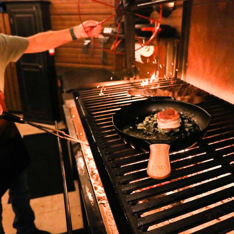 May include: A close-up of a cast iron skillet with a wooden handle, sitting on a black grill. The skillet contains a cooked steak with a bacon wrap and green onions. The grill is lit and has flames visible.