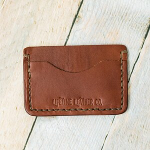 Puede incluir: Un porta tarjetas de cuero marr&oacute;n con un borde cosido y el texto "LIFETIME LEATHER CO." en relieve en la parte delantera.