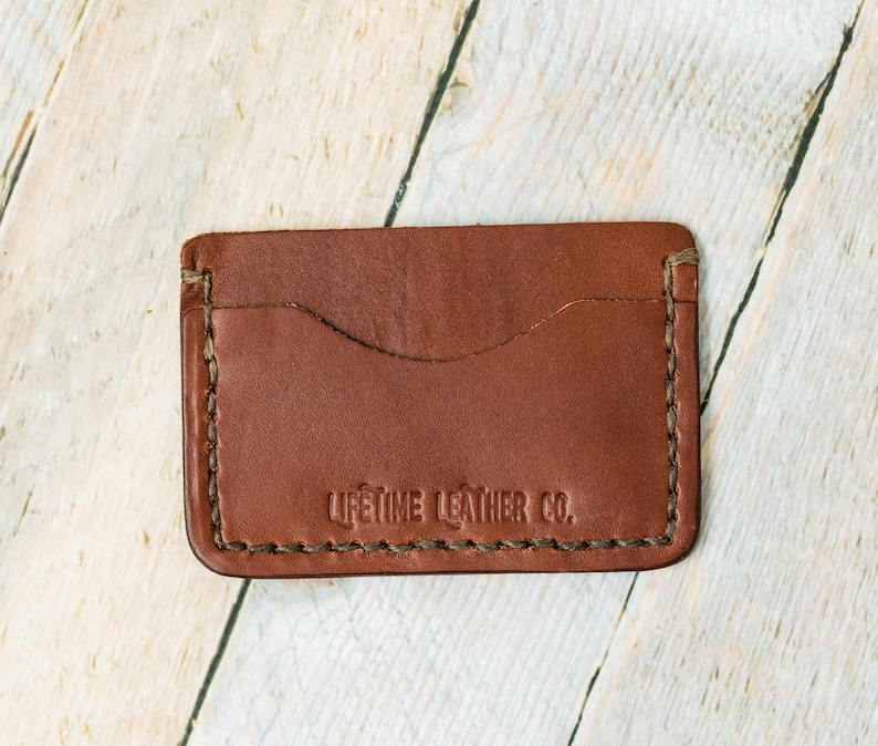 Puede incluir: Un porta tarjetas de cuero marr&oacute;n con un borde cosido y el texto "LIFETIME LEATHER CO." en relieve en la parte delantera.
