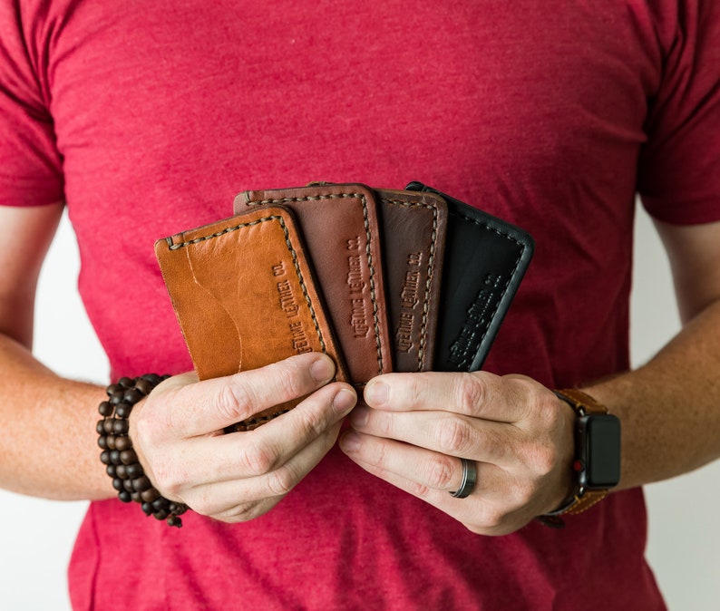 Puede incluir: Una persona sosteniendo cuatro porta tarjetas de cuero en diferentes colores. Los porta tarjetas est&aacute;n hechos por "Genuine Leather Co."