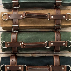 May include: Four rolled canvas tool rolls in various colors: olive green, brown, black, and dark green. Each roll features brown leather straps, silver metal buckles, and a leather handle for carrying. The rolls are arranged horizontally.